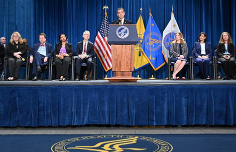 HHS Secretary Xavier Becerra speaks at a lectern while HRSA Administrator Carole Johnson and other HHS leaders are seated on stage.