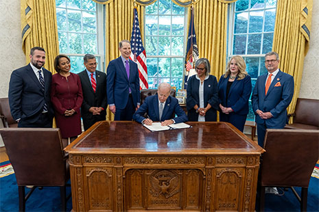 President Biden signs legislation at his desk in the Oval Office with attendees including Carole Johnson, Xavier Becerra, Ron Wyden, Robin Kelly, and patient advocates. 
