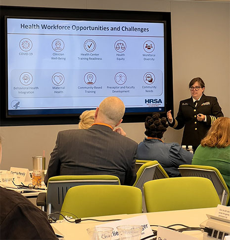 A woman gives a presentation in a conference room. A large screen on the wall displays information on health workforce opportunities and challenges.