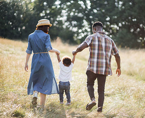 An adult woman and adult man hold a young child by the hand as they walk through a field.