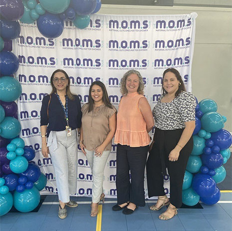 Four women pose in front of the M.O.M.S banner and colorful balloons.