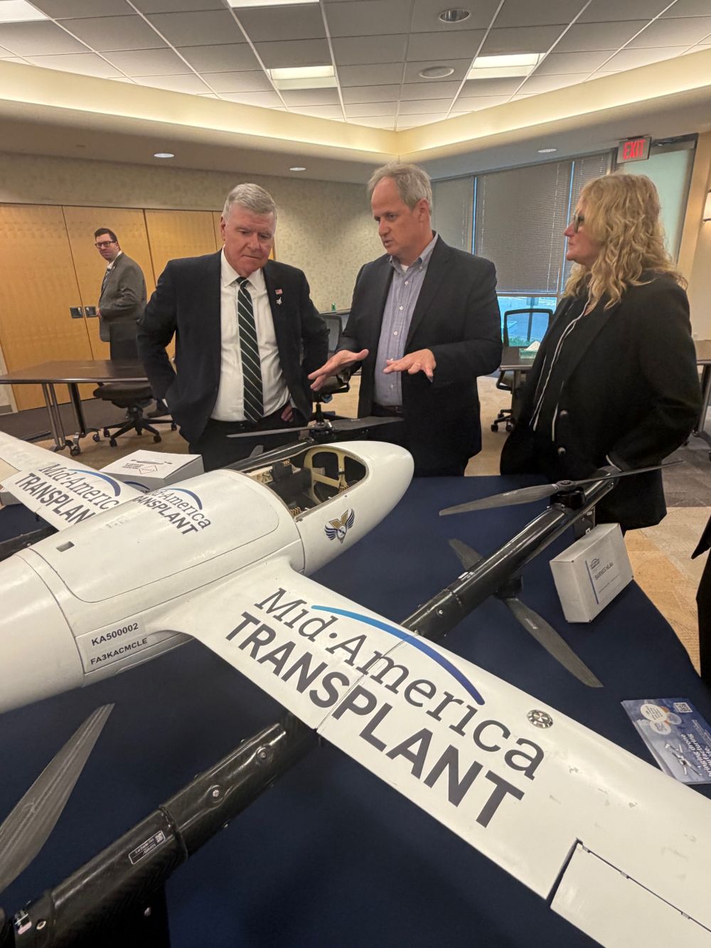 Three people are standing in front of a Mid-American Transplant Drone.