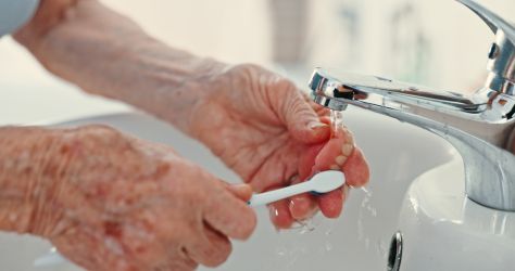 An older adult brushing their dentures with a toothbrush in the sink.