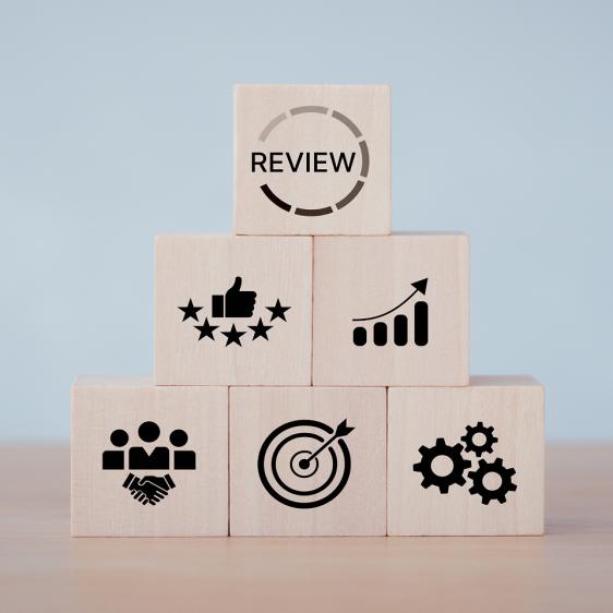 Pyramid of wooden blocks with icons; top block reads “REVIEW,” below icons for quality, growth, teamwork, targets, and process.