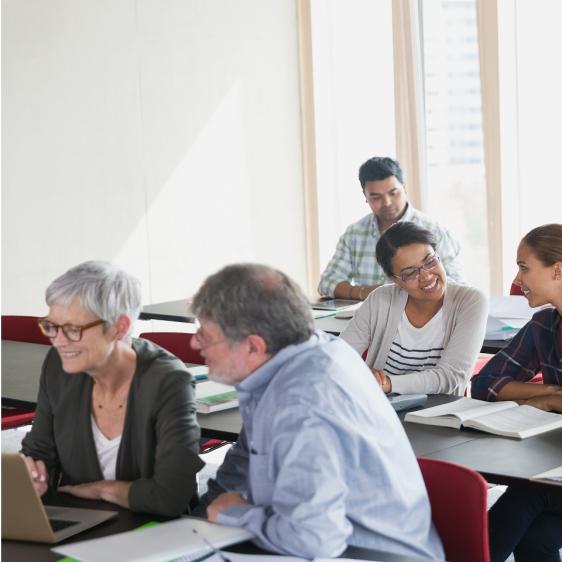 Adults seated at classroom tables during a training session, collaborating with laptops and open notebooks near large windows.