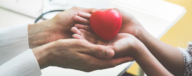 A doctor’s outstretched hands holding a young patient’s hands that contain a red heart. 