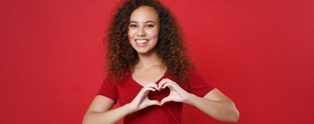 A woman wearing red making the shape of a heart with her hands. 